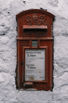 A vintage red postal box embedded in a white textured wall, featuring an emblem and the inscription 'G R' at the top. Below is a slot labeled 'Letters Only' and further down is a notice with collection times for mail from Monday to Saturday.
