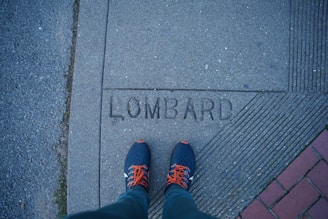 A pair of feet in blue sneakers with orange laces standing on a concrete sidewalk. The word 'LOMBARD' is engraved on the sidewalk. To the left, there's a gravel or asphalt surface and on the right, there are brick pavers.