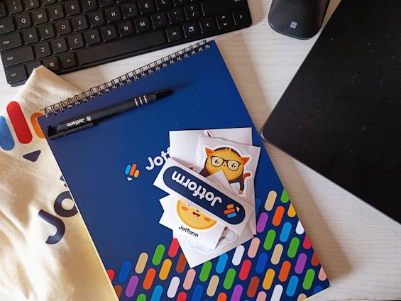 A workspace setting featuring a keyboard, a black pen, a spiral-bound notebook with a blue cover and colorful pattern, and a few playful stickers. The brand name 'Jotform' is visible on the pen, notebook, and stickers. A fabric surface and a black electronic device are also present.