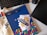A workspace setting featuring a keyboard, a black pen, a spiral-bound notebook with a blue cover and colorful pattern, and a few playful stickers. The brand name 'Jotform' is visible on the pen, notebook, and stickers. A fabric surface and a black electronic device are also present.