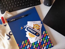 A workspace setting featuring a keyboard, a black pen, a spiral-bound notebook with a blue cover and colorful pattern, and a few playful stickers. The brand name 'Jotform' is visible on the pen, notebook, and stickers. A fabric surface and a black electronic device are also present.