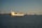 A large yellow cargo ship labeled 'Stolt Tankers' is prominent in the foreground, sailing on a calm sea. Several other cargo ships are visible in the background. A long suspension bridge spans across the horizon, below a clear sky. City buildings can be seen along the distant shoreline.