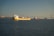 A large yellow cargo ship labeled 'Stolt Tankers' is prominent in the foreground, sailing on a calm sea. Several other cargo ships are visible in the background. A long suspension bridge spans across the horizon, below a clear sky. City buildings can be seen along the distant shoreline.