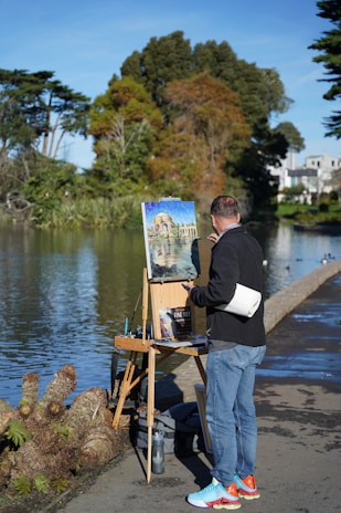 A man is painting a scenic view by a lake while standing on a path. He uses an easel and various painting supplies are visible, such as paintbrushes and a roll of paper towels. The painting depicts a building surrounded by trees and water. The surroundings are lush with greenery, and there are large trees around the serene lake.