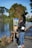 A man is painting a scenic view by a lake while standing on a path. He uses an easel and various painting supplies are visible, such as paintbrushes and a roll of paper towels. The painting depicts a building surrounded by trees and water. The surroundings are lush with greenery, and there are large trees around the serene lake.