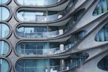 Curved modern architectural facade with a series of uniquely shaped balconies. The building features large glass panels and steel accents, creating a sleek and futuristic appearance. Each balcony is equipped with minimalistic white furniture, giving a sense of elegance and style.