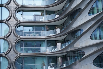 Curved modern architectural facade with a series of uniquely shaped balconies. The building features large glass panels and steel accents, creating a sleek and futuristic appearance. Each balcony is equipped with minimalistic white furniture, giving a sense of elegance and style.