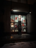 A storefront display featuring a large sculpted head on a pedestal. The windows are adorned with red silhouettes of martial artists and various small artifacts or decorative items. Photographic portraits are lined at the bottom of the window display. Dim lighting with shadows creates a mysterious ambiance.