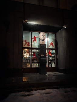 A storefront display featuring a large sculpted head on a pedestal. The windows are adorned with red silhouettes of martial artists and various small artifacts or decorative items. Photographic portraits are lined at the bottom of the window display. Dim lighting with shadows creates a mysterious ambiance.
