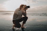 A person crouches on a reflective wet surface, holding a camera up to their face, capturing a photograph. The background reveals a serene, expansive seascape with soft, glowing sunlight filtering through scattered clouds, casting a gentle reflection on the water.