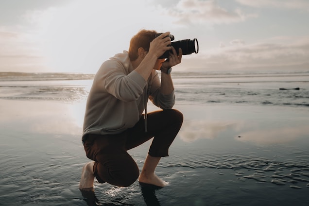 A person crouches on a reflective wet surface, holding a camera up to their face, capturing a photograph. The background reveals a serene, expansive seascape with soft, glowing sunlight filtering through scattered clouds, casting a gentle reflection on the water.