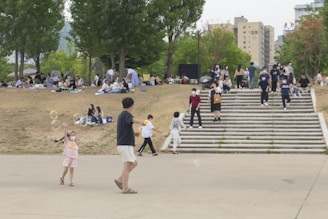 People are gathered in a park setting with children playing with bubbles. Groups are sitting on blankets on a grassy area, while others walk up and down wide steps. Trees provide some greenery, and there are buildings in the background.