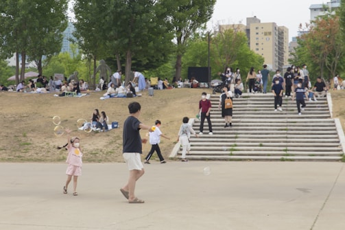 People are gathered in a park setting with children playing with bubbles. Groups are sitting on blankets on a grassy area, while others walk up and down wide steps. Trees provide some greenery, and there are buildings in the background.