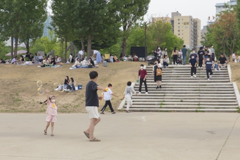 People are gathered in a park setting with children playing with bubbles. Groups are sitting on blankets on a grassy area, while others walk up and down wide steps. Trees provide some greenery, and there are buildings in the background.