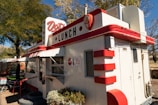 A small, retro-style diner with red and white accents. The building has signage indicating 'Dot's Lunch' and features classic diner elements such as rounded corners and decorative detailing. There is a window for ordering and outdoor seating is suggested by the nearby furniture. The setting is surrounded by trees, suggesting a pleasant outdoor atmosphere.