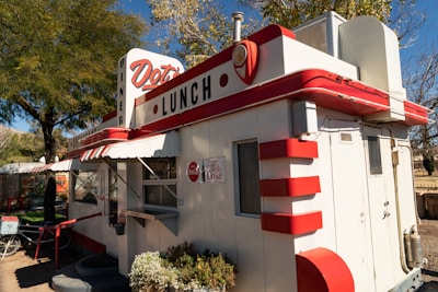 A small, retro-style diner with red and white accents. The building has signage indicating 'Dot's Lunch' and features classic diner elements such as rounded corners and decorative detailing. There is a window for ordering and outdoor seating is suggested by the nearby furniture. The setting is surrounded by trees, suggesting a pleasant outdoor atmosphere.