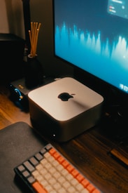 A compact Apple computer is placed on a wooden desk next to a mechanical keyboard with red, white, and black keys. A monitor displaying a waveform graphic sits to the right of the computer. There is also a black diffuser with sticks and an audio interface on the desk.
