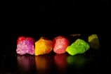 Vibrantly colored rocks are arranged on a reflective surface against a dark background, showcasing hues of purple, yellow, red, and green. The textures of the rocks are highlighted by the lighting, giving a crystalline appearance.