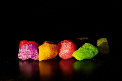 Vibrantly colored rocks are arranged on a reflective surface against a dark background, showcasing hues of purple, yellow, red, and green. The textures of the rocks are highlighted by the lighting, giving a crystalline appearance.