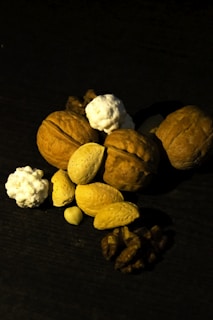 A collection of nuts including walnuts, almonds, and possibly some small popcorn-like elements are arranged on a dark surface. The lighting casts deep shadows, highlighting the textures of the nuts.