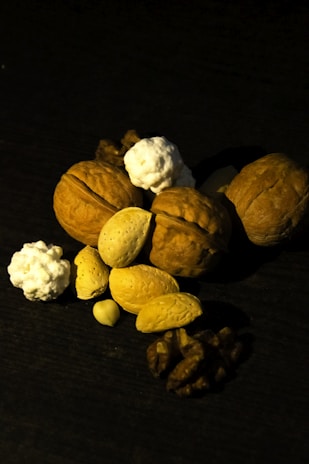A collection of nuts including walnuts, almonds, and possibly some small popcorn-like elements are arranged on a dark surface. The lighting casts deep shadows, highlighting the textures of the nuts.