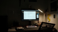 A dimly lit desk setup featuring a computer monitor displaying a document titled 'General Hardening Guideline'. The desk has a mechanical keyboard with blue and red keys, a lamp providing light on the right side, and various small items including notes pinned to the wall, a notebook, and a cup. There is a mesh office chair in front of the desk.