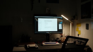 A dimly lit desk setup featuring a computer monitor displaying a document titled 'General Hardening Guideline'. The desk has a mechanical keyboard with blue and red keys, a lamp providing light on the right side, and various small items including notes pinned to the wall, a notebook, and a cup. There is a mesh office chair in front of the desk.