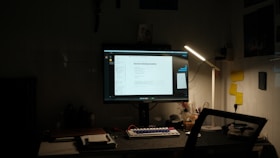 A dimly lit desk setup featuring a computer monitor displaying a document titled 'General Hardening Guideline'. The desk has a mechanical keyboard with blue and red keys, a lamp providing light on the right side, and various small items including notes pinned to the wall, a notebook, and a cup. There is a mesh office chair in front of the desk.