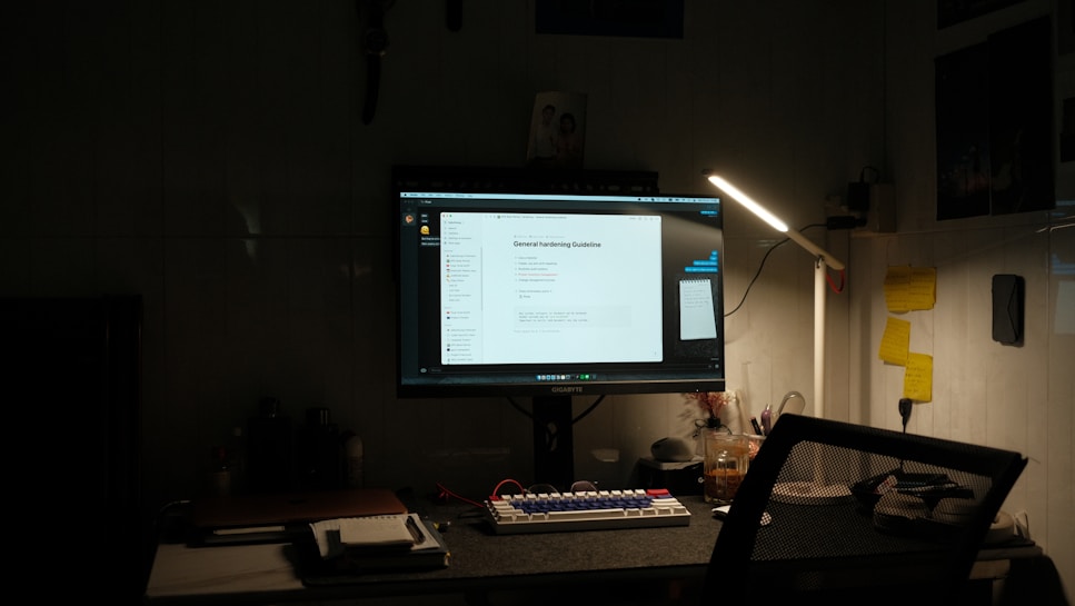 A dimly lit desk setup featuring a computer monitor displaying a document titled 'General Hardening Guideline'. The desk has a mechanical keyboard with blue and red keys, a lamp providing light on the right side, and various small items including notes pinned to the wall, a notebook, and a cup. There is a mesh office chair in front of the desk.