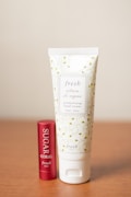 Two beauty products are displayed on a wooden surface: a tube of moisturizing hand cream with a decorative floral pattern and a cylinder of lip balm with a red metallic finish.