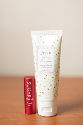 Two beauty products are displayed on a wooden surface: a tube of moisturizing hand cream with a decorative floral pattern and a cylinder of lip balm with a red metallic finish.