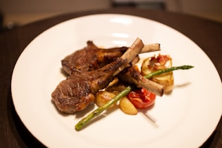 Grilled lamb chops are artfully arranged on a white plate, accompanied by roasted vegetables including asparagus, cherry tomato, and garlic cloves. A halved mushroom topped with crispy bacon adds texture and flavor. The food is presented in a fine dining style with a warm lighting accentuating the colors and textures.