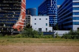 A cityscape featuring modern buildings with unique architectural designs. The foreground has a grassy area, while the background displays a variety of high-rise structures, including a red brick building with green terraces and a blue glass building with geometric patterns.