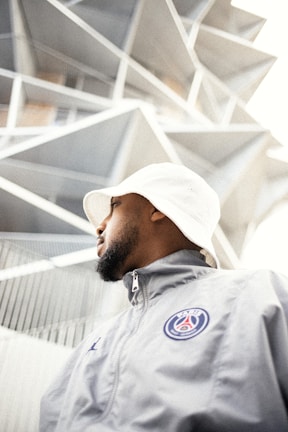 A person wearing a white bucket hat and light-grey jacket with a sports logo is gazing upwards. The background features a modern building with unique geometric architectural elements.