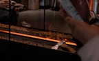 A modern fireplace design with flames running through a linear setup surrounded by small brown stones. An axe is placed on the edge and a person's hand is holding a smartphone nearby.