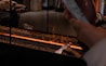 A modern fireplace design with flames running through a linear setup surrounded by small brown stones. An axe is placed on the edge and a person's hand is holding a smartphone nearby.