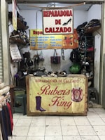 A small shoe repair shop with various signs advertising shoe repair services. The front table showcases a sign with the name 'Reparadora de Calzado Ruben's King' along with images of a boot and a lion. Shelves are stacked with shoes and shoe-related items. Tools and materials are visible, indicating a workspace for repairs.