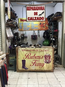 A small shoe repair shop with various signs advertising shoe repair services. The front table showcases a sign with the name 'Reparadora de Calzado Ruben's King' along with images of a boot and a lion. Shelves are stacked with shoes and shoe-related items. Tools and materials are visible, indicating a workspace for repairs.