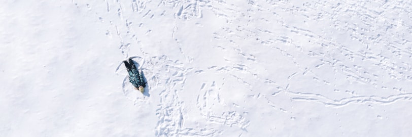 A single penguin stands on an expansive, snowy white surface. Its shadow is visible on the snow, and the area around the penguin appears to have multiple trails and markings, possibly from other penguins or movement through the snow.