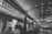 A bustling indoor market corridor with a high arched ceiling adorned with strings of lights. Several food stalls line the sides, including one prominently named 'Naan Stop Express'. Patrons are seen ordering and collecting food, adding to a lively and dynamic atmosphere.