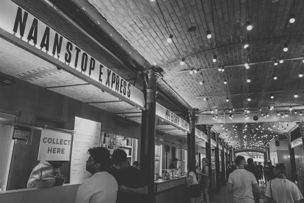 A bustling indoor market corridor with a high arched ceiling adorned with strings of lights. Several food stalls line the sides, including one prominently named 'Naan Stop Express'. Patrons are seen ordering and collecting food, adding to a lively and dynamic atmosphere.