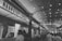 A bustling indoor market corridor with a high arched ceiling adorned with strings of lights. Several food stalls line the sides, including one prominently named 'Naan Stop Express'. Patrons are seen ordering and collecting food, adding to a lively and dynamic atmosphere.