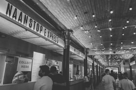 A bustling indoor market corridor with a high arched ceiling adorned with strings of lights. Several food stalls line the sides, including one prominently named 'Naan Stop Express'. Patrons are seen ordering and collecting food, adding to a lively and dynamic atmosphere.