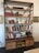 A large wooden bookshelf filled with a variety of books, some stacked vertically, others horizontally. On the shelves are decorative items including vases with white flowers, glassware, and a small statue. The shelves are supported by a metal frame with an industrial look. Below the shelves, there are several drawers and open spaces containing board games and other items.