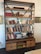 A large wooden bookshelf filled with a variety of books, some stacked vertically, others horizontally. On the shelves are decorative items including vases with white flowers, glassware, and a small statue. The shelves are supported by a metal frame with an industrial look. Below the shelves, there are several drawers and open spaces containing board games and other items.