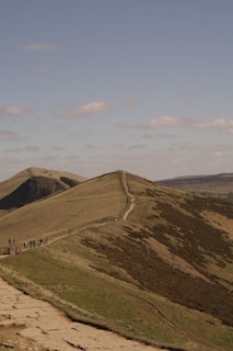 A scenic landscape depicting a winding trail along the crest of a grassy ridge. The path is bordered by a wooden fence and is frequented by several hikers. The background features rolling hills under a clear blue sky with scattered clouds.