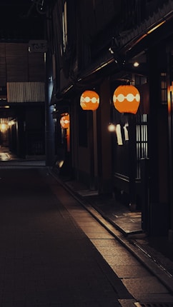 A dimly lit narrow street with traditional Japanese lanterns illuminating the dark pathway. The scene is quiet and atmospheric, with warm orange lights contrasting against the dark surroundings. The architecture features wooden and paper elements typical of old Japanese buildings.