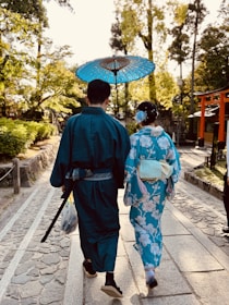 A couple dressed in traditional Japanese attire walks down a stone path in a garden-like setting. The man is carrying a sword and the woman holds a blue parasol. They are surrounded by lush greenery and traditional architecture.