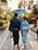 A couple dressed in traditional Japanese attire walks down a stone path in a garden-like setting. The man is carrying a sword and the woman holds a blue parasol. They are surrounded by lush greenery and traditional architecture.