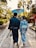 A couple dressed in traditional Japanese attire walks down a stone path in a garden-like setting. The man is carrying a sword and the woman holds a blue parasol. They are surrounded by lush greenery and traditional architecture.